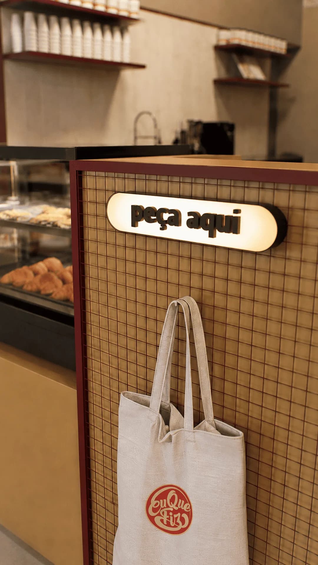 Balcão de pedidos com letreiro iluminado "Peça Aqui", sacola de pano da marca e vitrine de croissants ao fundo.
