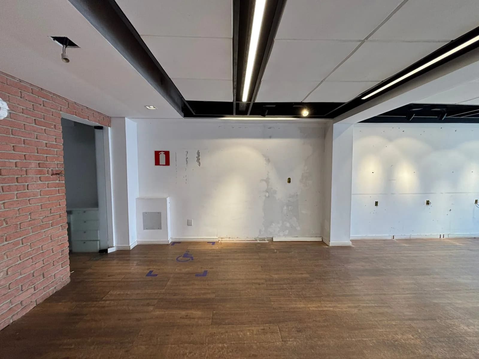 Antes da Gigi Bambini: salão vazio visto de lado, com parede de tijolos à esquerda e forro com luminárias lineares.