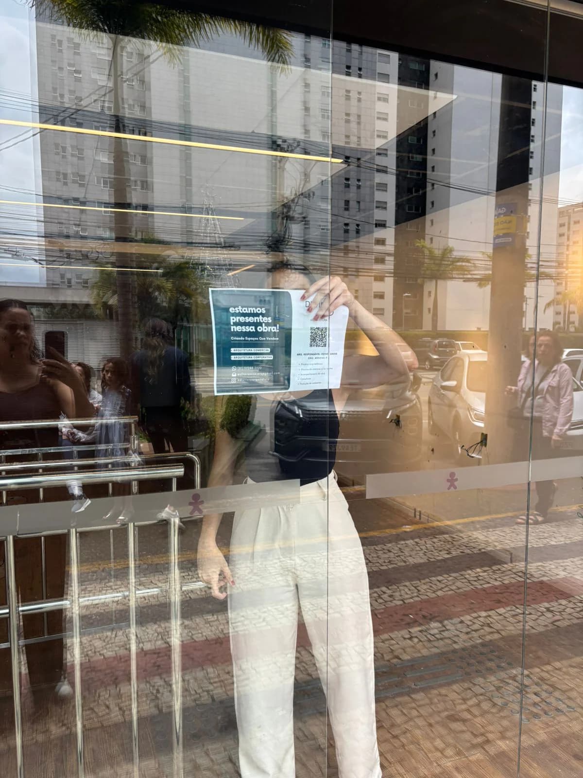 Vitrine de loja com o reflexo da rua no vidro e uma pessoa segurando um cartaz do lado de dentro.